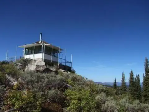 A fire lookout tower sits atop a mountain ridge surrounded by trees and clear blue sky.