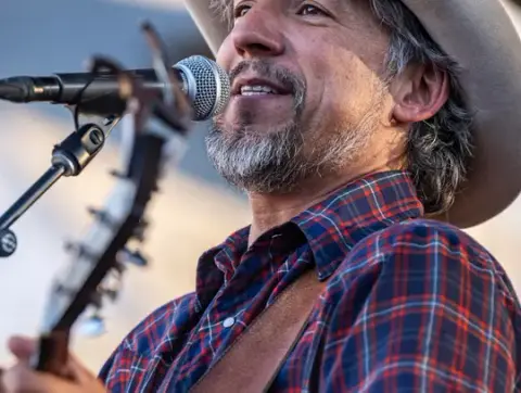 A man wearing a cowboy hat sings into a microphone while holding a guitar.