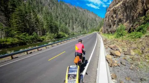 A cyclist with a trailer rides along a winding road through a mountainous landscape.