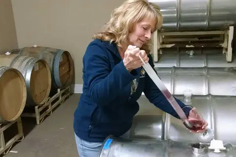 A woman uses a thief to sample wine from a metal barrel in a cellar.