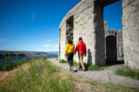 A couple walks along a path next to a stone structure overlooking water and a landscape.