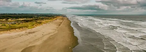 An aerial view of a long sandy beach with waves rolling in under a cloudy sky.