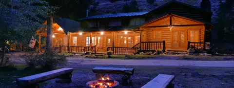 A brightly lit log cabin exterior at dusk with a campfire pit in the foreground.