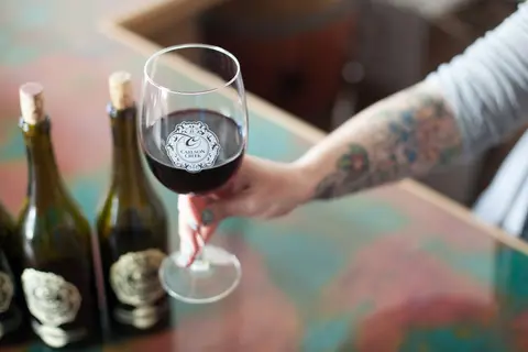 A person holds a glass of red wine next to wine bottles on a table.