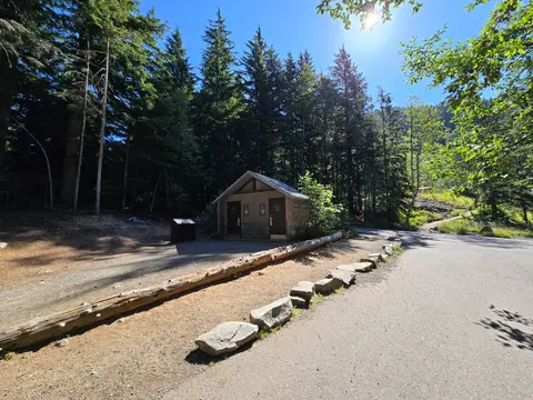 A small restroom building stands near a paved parking area surrounded by evergreen trees.