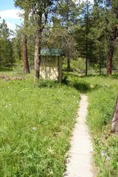 A small outhouse stands at the end of a path in a grassy wooded area.