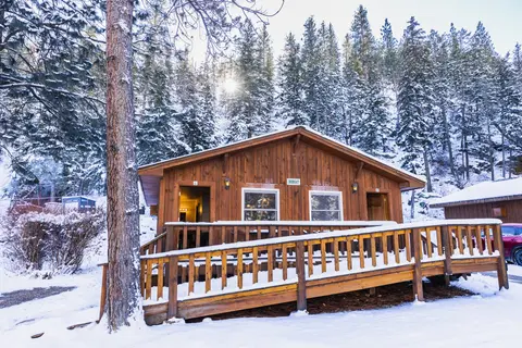 A wooden cabin in a snowy forest with a ramp leading to the porch.