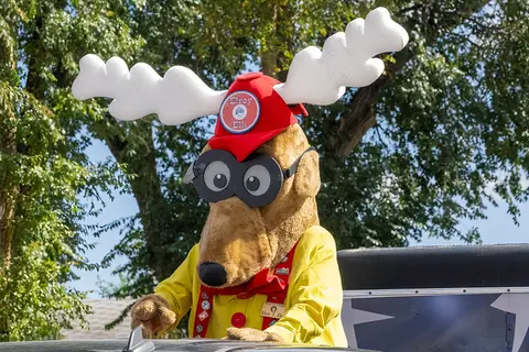 A mascot character wearing a red hat and white antlers rides on a vehicle.
