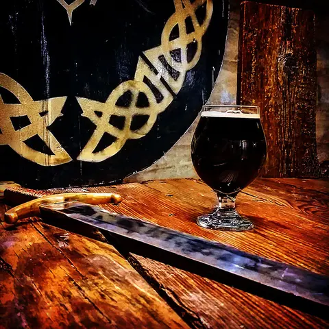A glass of dark beverage on a wooden table with a sword and a decorative shield.
