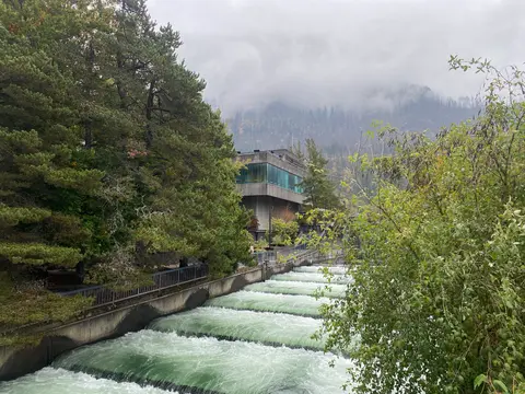 A modern building sits next to a tiered water channel surrounded by lush trees and foggy mountains.