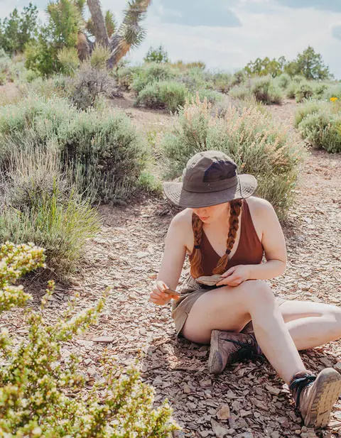 A person sits on the ground in a desert landscape, examining rocks.