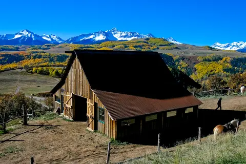 A rustic wooden barn stands in a landscape with snow-capped mountains in the background.