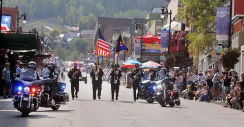A parade procession with an honor guard and police motorcycles moves down a street lined with buildings and crowds.