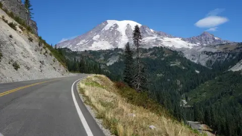 A winding road curves through a mountainous landscape with a snow-capped peak in the background.