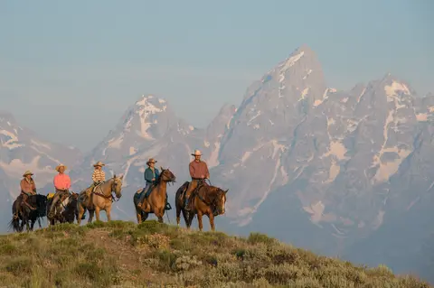 A group of horseback riders ride along a ridge with mountains in the background.