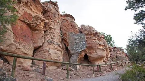 A dirt path runs alongside a wooden fence beneath a rocky cliff formation.