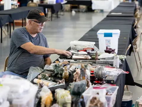 A man sits at a table covered in crystals and minerals inside a large hall.