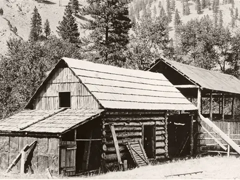 Black and white historic photo showing rustic log buildings with shingle roofs.