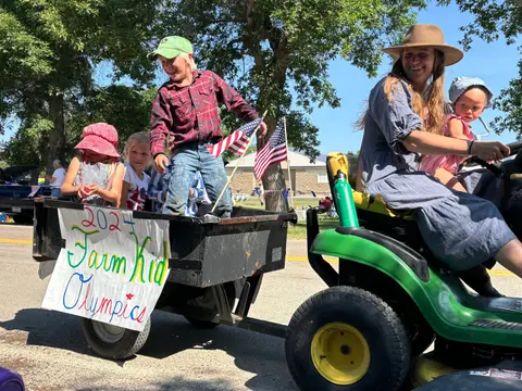 People riding on a small trailer attached to a lawn tractor in a parade.