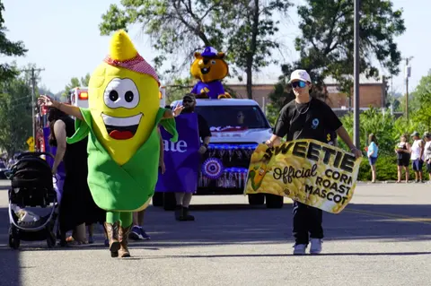 A large corn mascot walks down the street in a parade.