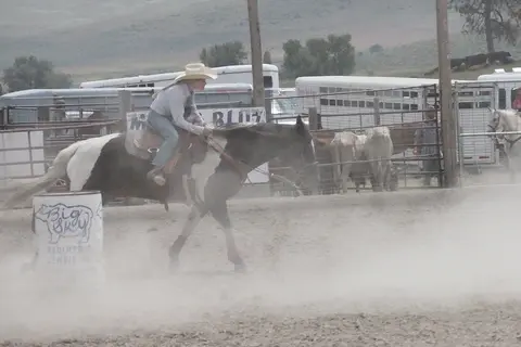 A cowgirl rides a pinto horse in a dusty arena with a barrel.