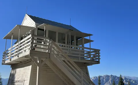 A white fire lookout tower stands against a clear blue sky with mountains in the background.
