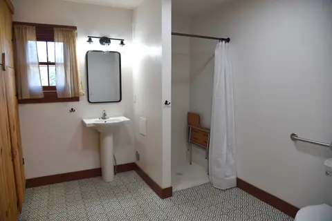 An interior view of a modern accessible bathroom with a pedestal sink, shower with a bench, and patterned tile flooring.