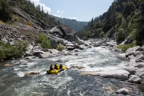 A yellow raft navigates a rocky river surrounded by mountains and trees.