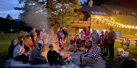 A large group of people gather around an outdoor campfire at dusk.