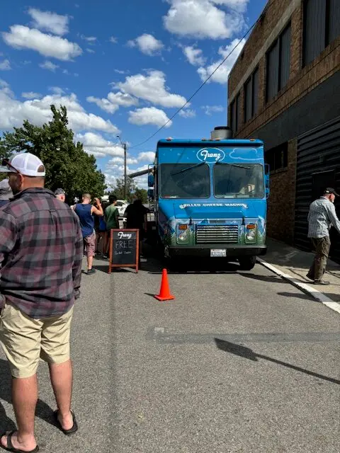 People stand in line next to a blue food truck on an asphalt street.