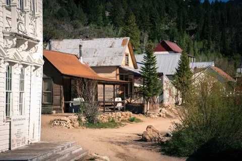 Exterior view of historic wooden buildings along a dirt street with a forest behind.