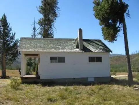 An old white building with a porch stands in a grassy area surrounded by trees.