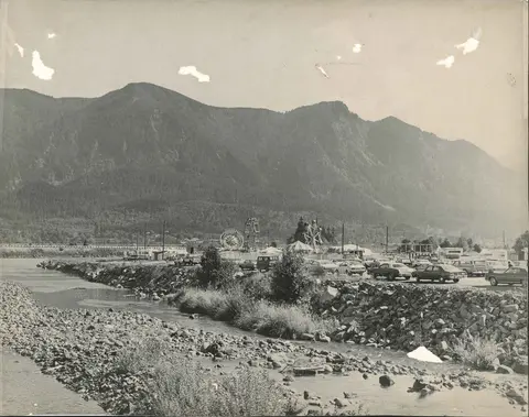 A vintage black and white photograph shows a river with a carnival and cars parked along the bank in front of mountains.