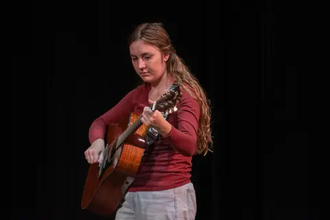 A young woman plays an acoustic guitar on stage.