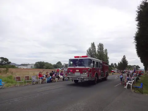 A fire truck drives down a road past a crowd of spectators sitting in chairs.
