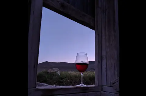 A glass of red wine sits on a wooden window sill overlooking a landscape at dusk.
