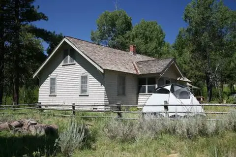 A light-colored wooden house stands behind a rustic wooden fence with a tent in the yard.