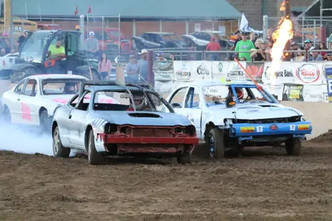 Several damaged cars participate in a demolition derby event on a dirt track.