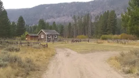 A dirt road leads to a couple of log cabins surrounded by fences in a valley with mountains and trees in the background.