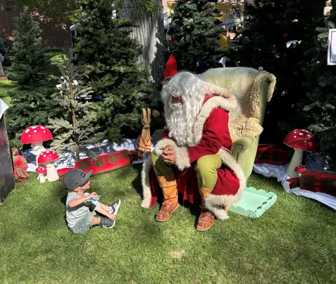 A young child sits on the grass talking to a person dressed as Santa Claus.