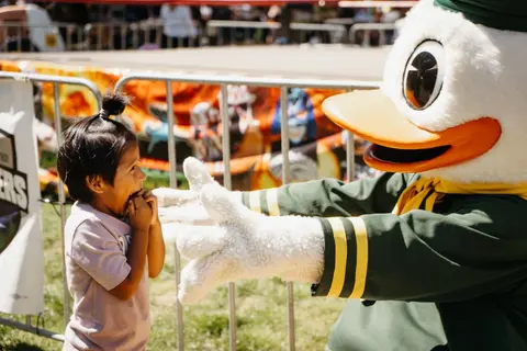 A child excitedly looks at a costumed duck mascot behind a metal barrier.