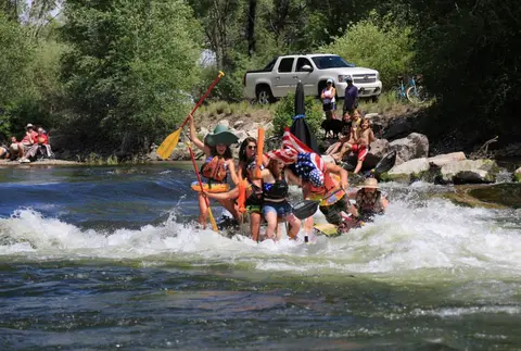 People on a raft navigating river rapids while spectators watch from the shore.