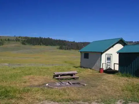 A small building with a green roof sits in a grassy field with a picnic table and fire ring.