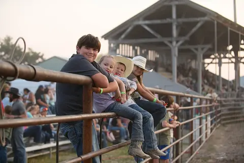 Several children sit on a metal fence watching an event near a grandstand.