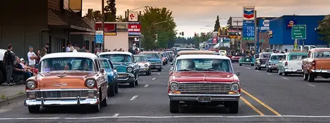 A line of vintage cars driving down a town street with spectators on the sidewalk.