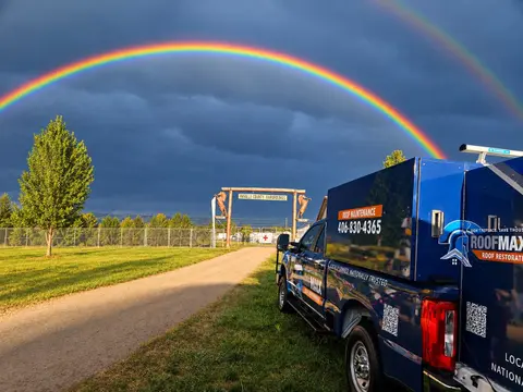 A double rainbow arches over a dirt road and a blue truck parked near an entrance gate.