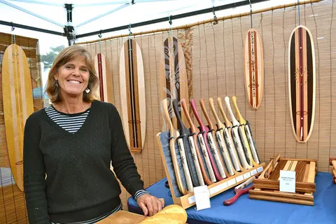 A woman smiles next to wooden crafts and surfboards at a market booth.