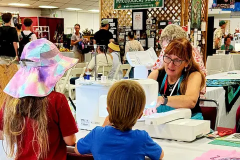 People gather indoors at a fair or convention where a woman helps children use sewing machines.