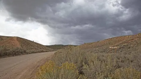 A dirt road winds through hilly desert landscape under a stormy sky.