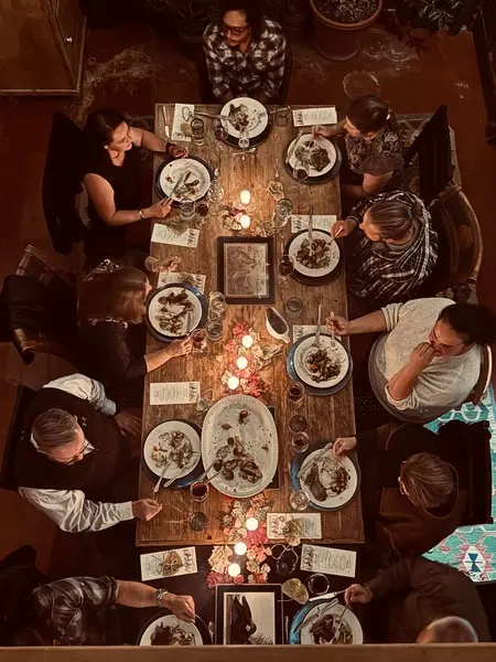 A high-angle view shows a group of people sitting around a long wooden dining table eating dinner by candlelight.